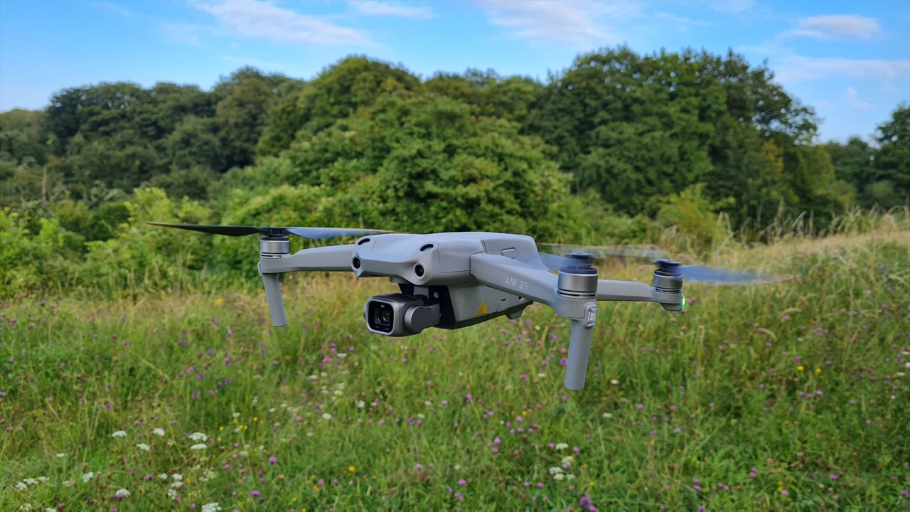 Dron compacto de cuatro hélices con su cámara al frente, en vuelo a baja altura sobre un potrero de pasto alto, con árboles al fondo en un día despejado